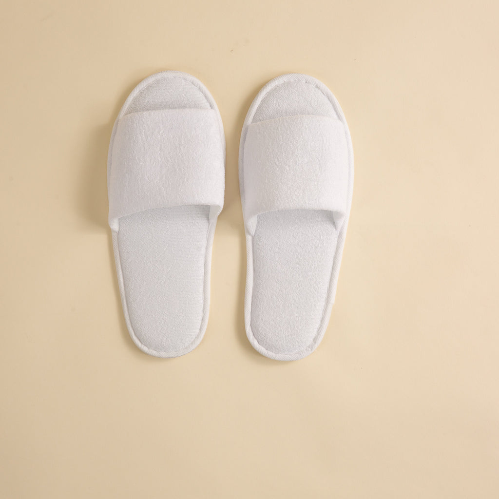 A pair of white towelling open-toe slippers placed on a neutral background.