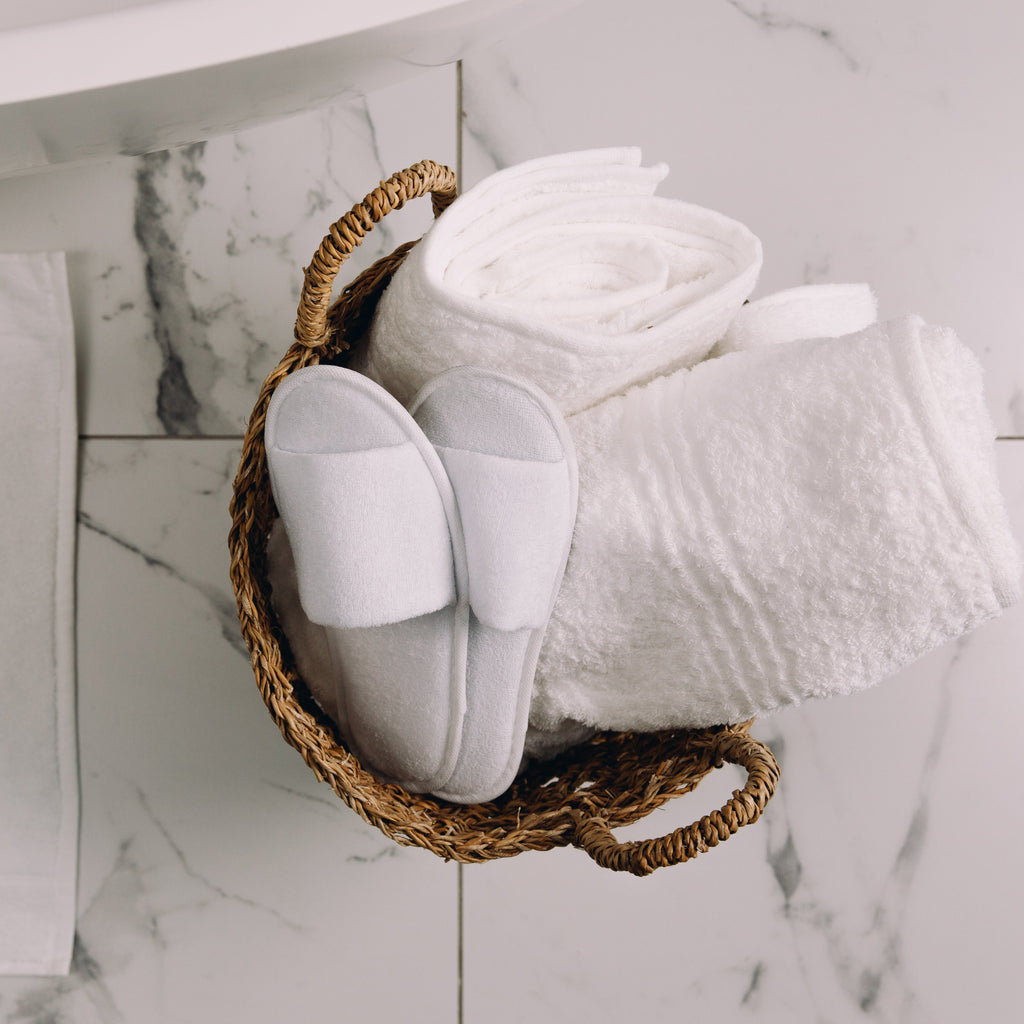 White slippers and folded towels in a woven basket on a marble floor.