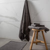 A set of luxury towels in sage, charcoal cloud, and white colors, displayed on a wooden stool and rack, with a decorative bathroom setting in the background featuring a marble wall and a dark grey rug.