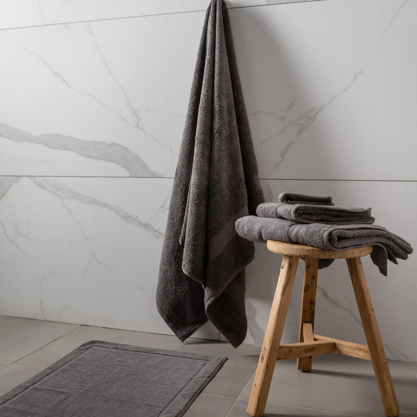 A set of luxury towels in sage, charcoal cloud, and white colors, displayed on a wooden stool and rack, with a decorative bathroom setting in the background featuring a marble wall and a dark grey rug.