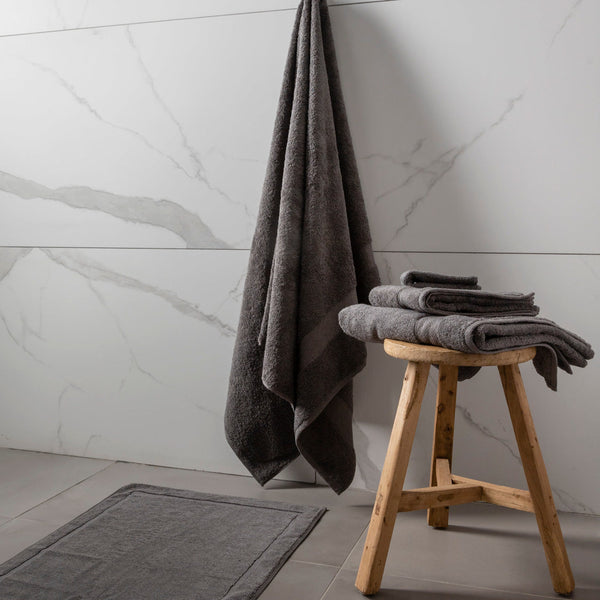 A set of bath towels in a bathroom setting, with one towel hanging on a wooden stool and the other two on a grey rug. The towels are in shades of grey and charcoal cloud, and the floor has a tiled design.