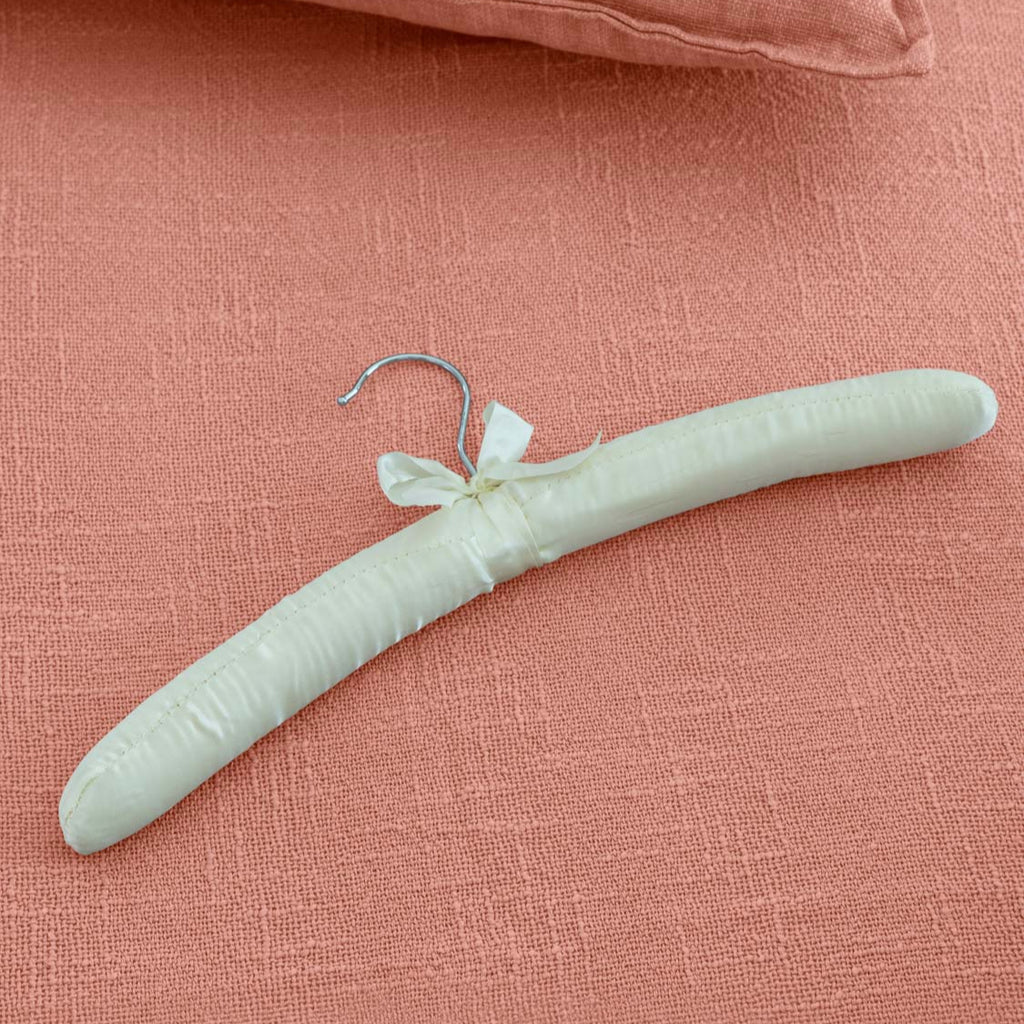 A satin hanger with a hook at one end, placed on a closet rod with a pink pillow and a satin-covered chair in the background.