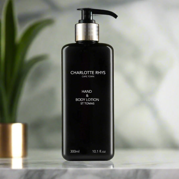 A black pump bottle of Charlotte Rhys Hand & Body Lotion with a label that reads 'Hand & Body Lotion, 300ml'. The bottle is placed on a marble surface with a plant in the background.