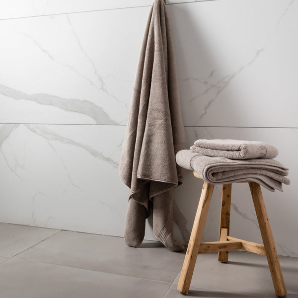 A set of folded towels in varying shades of gray and blue, placed on a wooden stool against a marble-tiled wall in a bathroom setting.